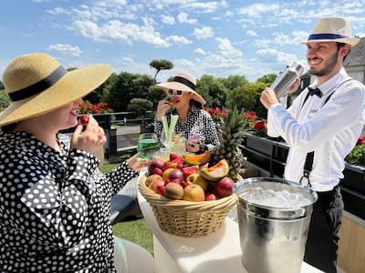 Two women in hats enjoy fruit, bartender prepares a cocktail, summer atmosphere.